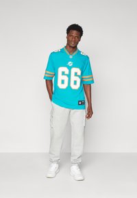 Ζέρσεϊ NFL Miami Dolphins σε aqua με πορτοκαλί ρίγες, με τον αριθμό 66, συνδυασμένο με γκρι cargo παντελόνια και λευκά αθλητικά παπούτσια.