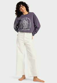 Gray long-sleeve t-shirt with a tiger graphic and text on the front, paired with white wide-leg pants, displayed against a neutral background.