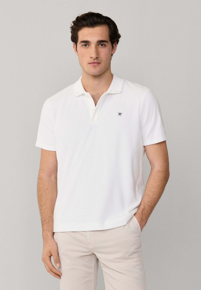 Giovane uomo con capelli scuri che indossa una polo bianca con un piccolo logo nero a forma di pagaie incrociate, pantaloni beige, in piedi contro uno sfondo semplice.