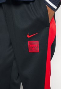 Pantalones deportivos negros con franjas laterales rojas, que presentan un logo rectangular de los Bulls en rojo y un pequeño swoosh de Nike en rojo. Fabricados con tejido suave.