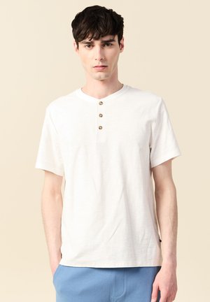 Giovane uomo che indossa una maglia henley bianca a maniche corte con tre bottoni in legno e pantaloni blu, su uno sfondo beige.