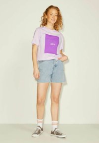 Camisa morada de manga corta con un gráfico cuadrado y texto blanco "JJXX" combinada con pantalones cortos de denim azul claro y zapatillas de lona grises.