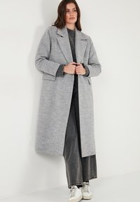 Grijze wollen jas met een lange, rechte snit, met klepjes en zijzakken, gecombineerd met donkere wijde broek en witte sneakers.