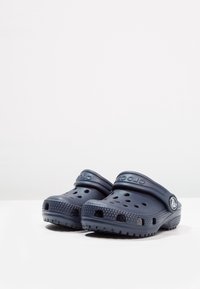 Para granatowych klapek Crocs z otworami wentylacyjnymi i regulowanymi paskami piętowymi na białej powierzchni.