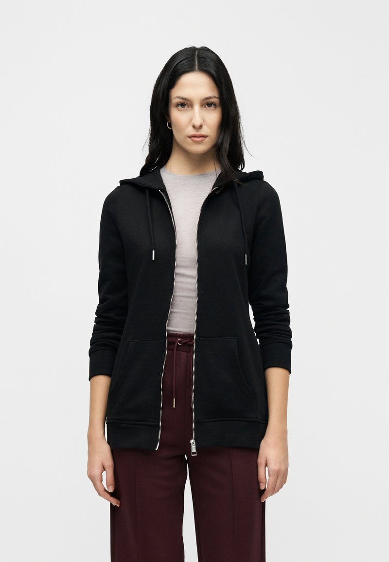 Black zip-up hoodie made of soft fabric, featuring a kangaroo pocket, drawstring hood, and ribbed cuffs. Paired with burgundy pants.