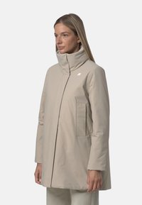 Beżowy długi płaszcz z wysokim kołnierzem, zamkiem z przodu i dwoma bocznymi kieszeniami. Posiada subtelne logo na piersi, wykonany z gładkiego, lekkiego materiału.