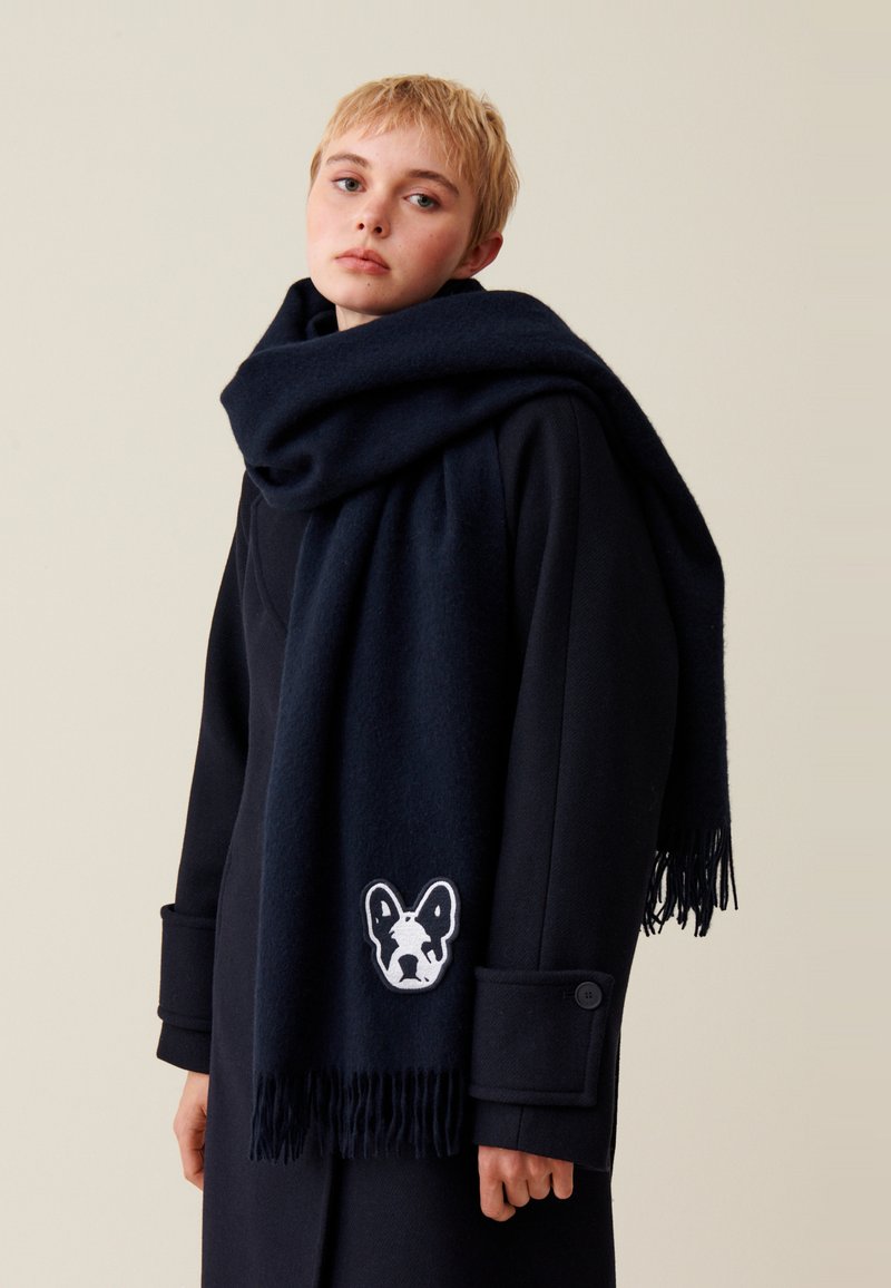 Écharpe en laine navy avec des bords frangés et un design de Bulldog Français brodé en blanc, portée sur un manteau sombre avec un grand détail de bouton.