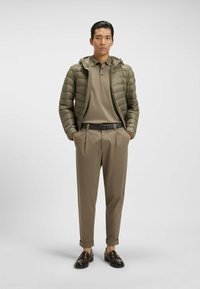 Kurtka puchowa w oliwkowym kolorze, zapinana na zamek, w połączeniu z beżową koszulą polo, beżowymi spodniami i brązowymi skórzanymi mokasynami. Minimalistyczny design.