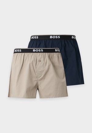 Homme torse nu debout pieds nus portant un boxer gris clair avec une ceinture noire BOSS sur un fond blanc uni.