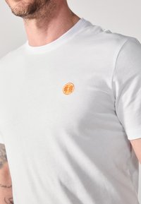 White short-sleeve t-shirt made from cotton, featuring an embroidered orange slice on the left chest area. Smooth texture, classic fit.