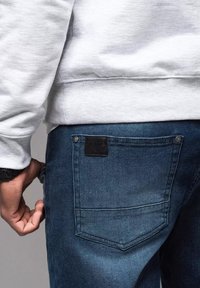 Jeans in denim blu scuro con tasca posteriore dotata di etichetta in pelle nera, abbinati a una felpa grigia chiara. Tessuto strutturato e vestibilità casual.