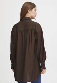 Brown long-sleeve shirt with a collared neckline, textured fabric, and pleated back detail, designed for a loose fit.
