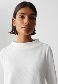 Vrouw met kort donker haar in een witte gebreide lange mouwen top, die recht vooruit kijkt met een neutrale uitdrukking tegen een effen achtergrond.