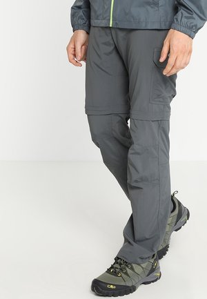 Hombre con pantalones cargo convertibles gris oscuro y zapatos de senderismo verde grisáceo, de pie con las piernas cruzadas.