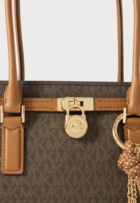 Borsa a spalla in tessuto marrone a fantasia con manici in pelle liscia color cammello. Presenta una chiusura a lucchetto in tonalità oro e una catena decorativa con un accento testurizzato.