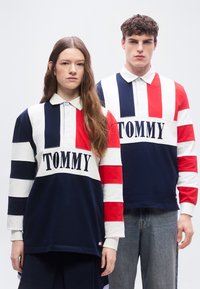 Νεαρός άνδρας και γυναίκα στέκονται δίπλα-δίπλα φορώντας ίδιες μακρυμάνικες ριγέ ρούγκμπι μπλούζες Tommy Hilfiger με σκούρο μπλε, λευκές και κόκκινες ρίγες.