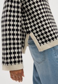 Maglione lavorato a maglia con un motivo a scacchi nero e crema, polsini a coste, fessure laterali e vestibilità rilassata, abbinato a jeans blu chiaro.