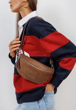 Kobieta w czerwono-granatowej w paski bluzie z długim rękawem i jasnych dżinsach, niosąca na ramieniu brązową torbę typu crossbody z zamszu i skóry.