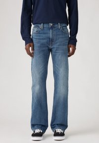 Jeans de denim azul claro con un ajuste relajado, decoloración sutil, diseño de cinco bolsillos y detalles de costura oscuros. Combinados con zapatillas negras.