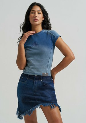 Femme aux longs cheveux foncés portant un t-shirt ajusté dégradé bleu et une jupe en denim bleu foncé à ourlet effiloché, posant devant un fond uni.