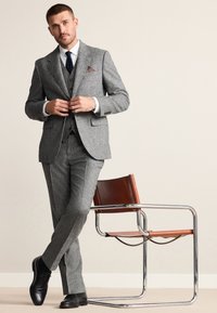 Costume en tweed gris avec pantalon assorti, chemise blanche, cravate bleu marine et pochette. Chaussures habillées noires. Fauteuil en cuir marron avec pieds en chrome.