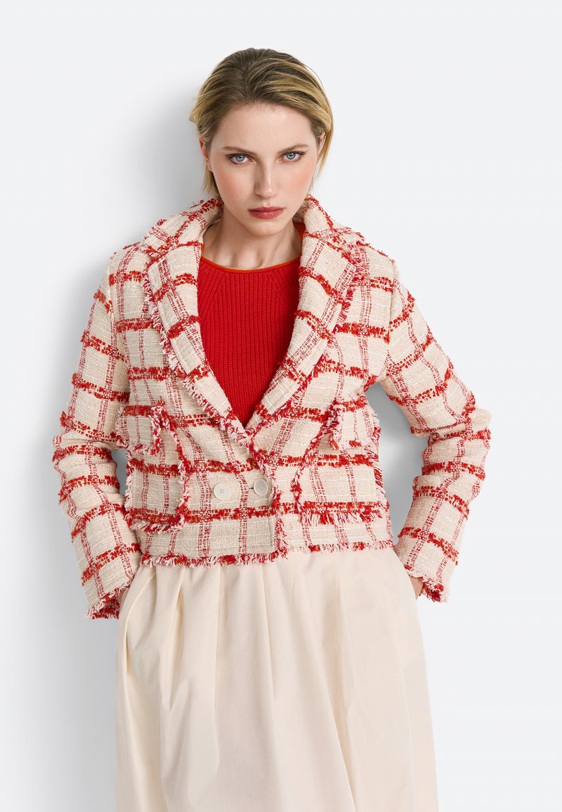 Veste en tweed à carreaux rouges et crème avec une coupe ajustée, des revers larges et deux boutons. Portée sur un top en tricot rouge, associée à une jupe crème.