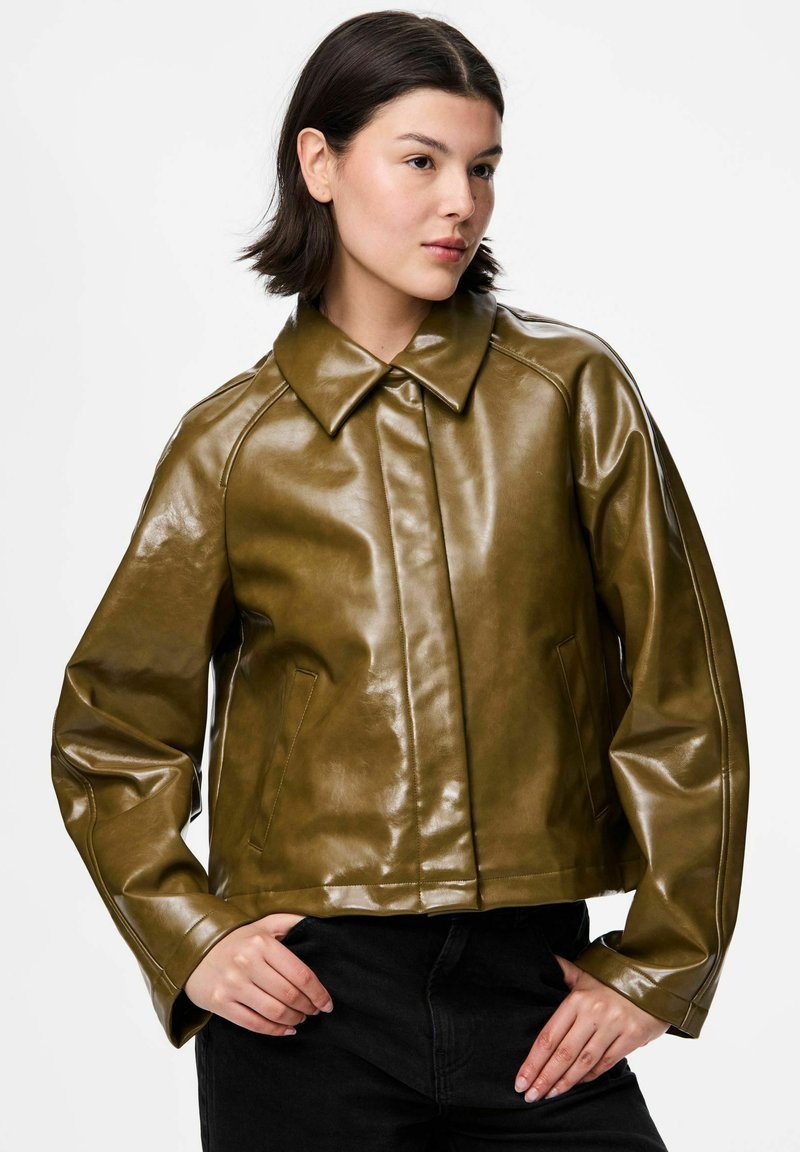 Sieviete, tērpusies ar īsu oliņu zaļu ādas jaku ar priekšējām kabatām un melniem bikšiem, rokas atspiedušās uz gurniem, skatās nedaudz pa labi.