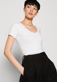 T-shirt ajusté blanc avec un col en V et des manches courtes, associé à un pantalon noir texturé taille haute. Design simple, tissu lisse.