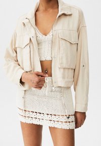 Beige cropped jacket with two front pockets, paired with a white crochet top and matching skirt featuring a textured pattern and drawstring detail.