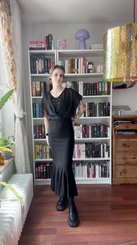 Melna kleita ar plūstošām piedurknēm un viļņotu apakšmalu, saskaņota ar masīvām melnām zābakiem. Geometrisks raksts uz augšdaļas. Grāmatu plaukts fona.