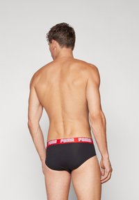 Sorte trusser med et rødt elastisk taljebånd, der har hvid "PUMA" branding. Stoffet ser glat ud, med en tætsiddende design.