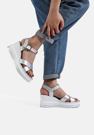 Sandali wedge argentati con cinghie metalliche incrociate, fibbia alla caviglia e suola piatta bianca. Indossati con jeans di denim arrotolati.