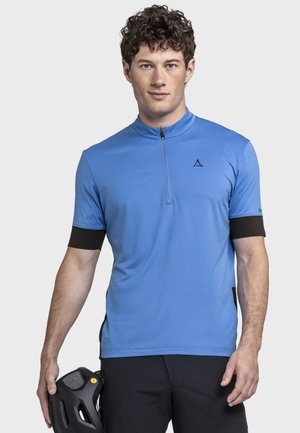 Mann mit lockigem Haar, der ein blaues kurzärmliges Radsporttrikot trägt und in seiner linken Hand einen schwarzen Fahrradhelm hält.