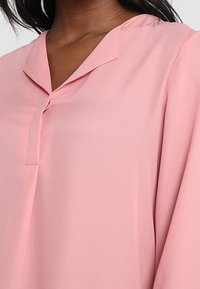 Blouse rose clair en tissu lisse, avec un col en V et une patte de boutonnage à l'avant, dotée de manches amples.