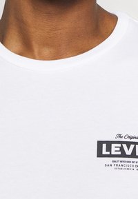 White cotton t-shirt featuring a round neck, ribbed collar, and black printed logo with text on the front, positioned on the left side.