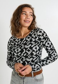Haut à manches longues en tissu noir avec un motif abstrait blanc. Présente un col rond et une coupe slim, associé à un jean clair taille haute.