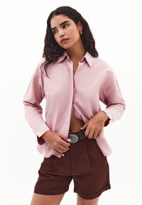 Lyserød skjorte med lange ærmer, krave og knapper, parret med brune plisserede shorts. Har et sølvfarvet bælte med en dekorativ spænde.