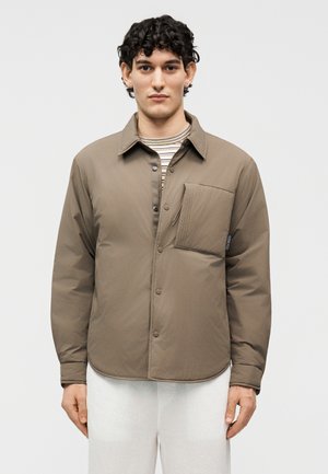 Muž s krátkymi kučeravými vlasmi, oblečený v taupe bundu na gombíky cez pruhované tričko a biele nohavice, stojaci pred jednofarebným pozadím.