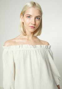 Top branco de ombro descoberto, feito de tecido texturizado, apresentando um decote franzido e mangas longas de corte solto com pregas subtis.