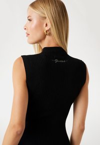 Kobieta z długimi blond włosami, ubrana w bezrękawnik z czarnego prążkowanego materiału z logo "Guess" na plecach oraz dużymi złotymi kolczykami, zwrócona w lewo.
