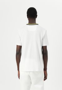 Polo shirt blanc à manches courtes avec un col côtelé noir et jaune, au design minimaliste et à la texture en coton lisse.