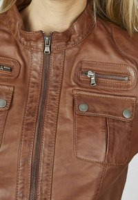 Giacca in pelle marrone con colletto alto, zip frontale, due tasche sul petto con zip e bottoni decorativi. Texture liscia con dettagli di cucitura.