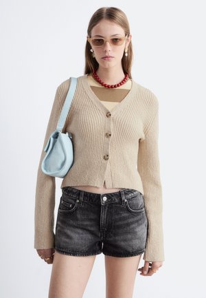Jeune femme portant un cardigan côtelé beige, un short en jean noir, un collier de perles rouges, des lunettes de soleil beiges, et portant un sac à épaule bleu clair.
