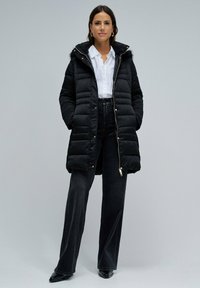 Abrigo acolchado negro con capucha forrada de pelo, cremallera frontal y detalles en dorado. Llevado sobre una camisa blanca y combinado con vaqueros oscuros.