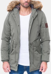 Parka verde con cappuccio foderato in pelliccia, vita con cordoncino, tasche frontali e una toppa in pelle marrone sull manica.