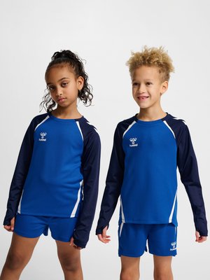 To børn i blå og marineblåt sportstøj, stående side om side, den ene med krøllet hår og smilende, den anden med samlet hår og ser seriøs ud.