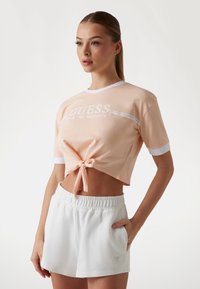 Persikofärgad kortärmad T-shirt med vita detaljer och knut framtill, ihopparad med vita shorts med elastisk midja och en logotyp.