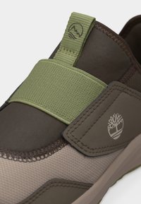 Κοντινή λήψη καφέ και μπεζ αθλητικού παπουτσιού με πράσινο ελαστικό λουράκι και ταινία για τράβηγμα, με λευκό λογότυπο δέντρου στην πλευρική επιφάνεια.