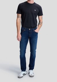Homme portant un jean bleu foncé, un t-shirt Lee noir et des baskets blanches, debout avec une main dans la poche devant un fond gris.