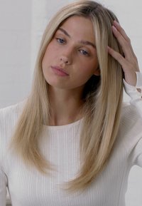 Lunghi capelli biondi lisci parzialmente nascosti dietro l'orecchio, indossa una maglietta bianca a maniche lunghe con coste, con una pelle liscia e un'espressione neutra.